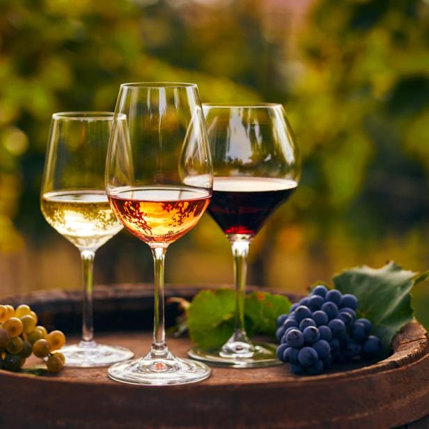 Drei Gläser mit Weißwein, Rosé und Rotwein stehen auf einem alten Holzfass mit Weintrauben.
