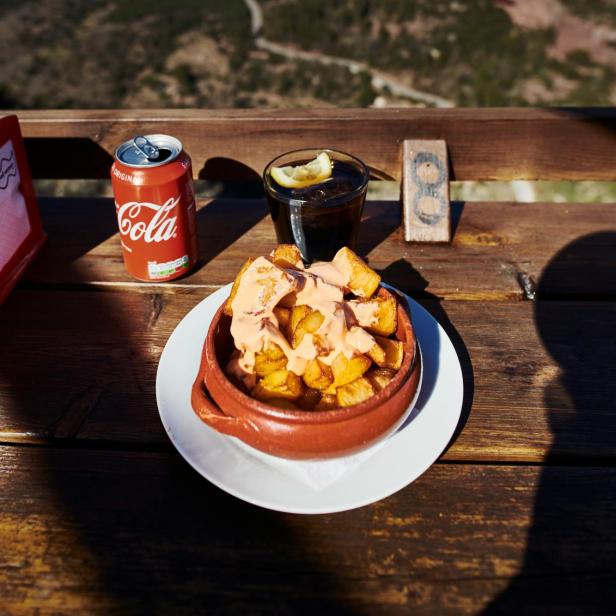 Patatas Bravas, Cola und ein Getränk mit Zitrone vor einer Berglandschaft.