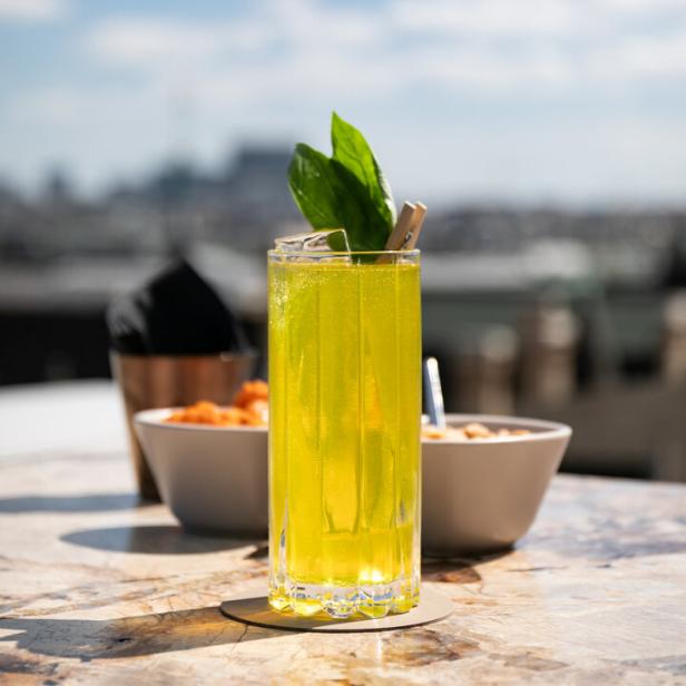 Ein gelber Cocktail mit Basilikumgarnitur auf einem Tisch im Freien serviert.