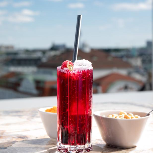 Ein roter Cocktail mit Himbeere und einem Metallstrohhalm auf einem Tisch mit Snacks.