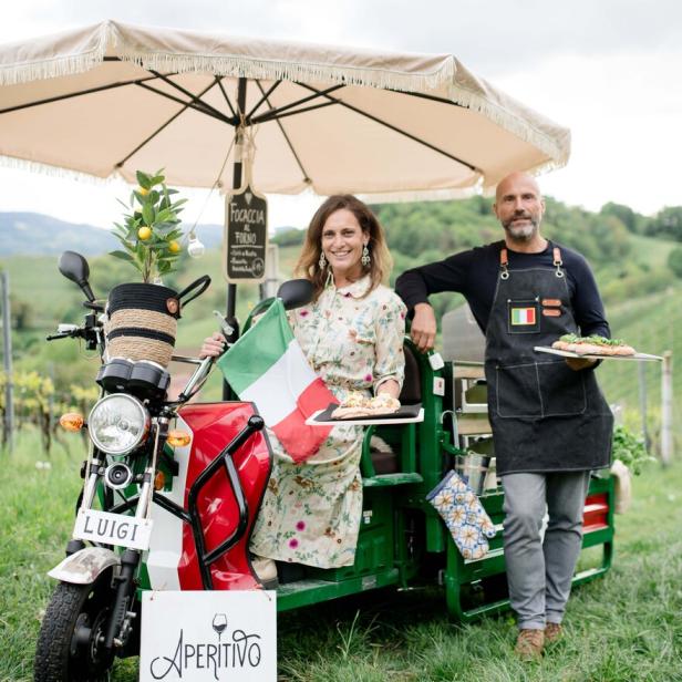 Eine Frau mit Italienischer Flagge und ein Mann mit Pizza stehen vor einem „Aperitivo“-Mobil.