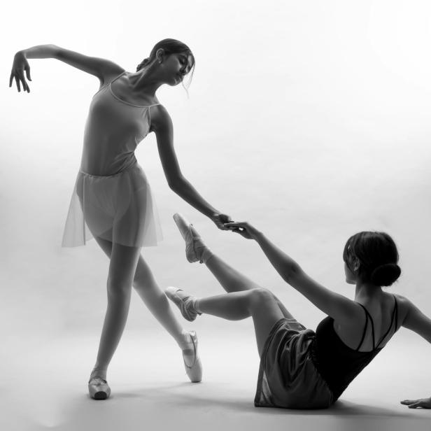 Eine stehende Ballerina reicht einer sitzenden Ballerina die Hand.