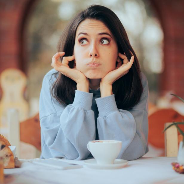 Eine gelangweilte Frau sitzt an einem Tisch mit einer Tasse Kaffee.