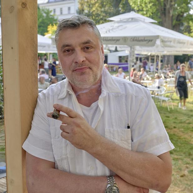Ein Mann lehnt an einem Holzpfosten und raucht eine Zigarre. Im Hintergrund ein Biergarten.