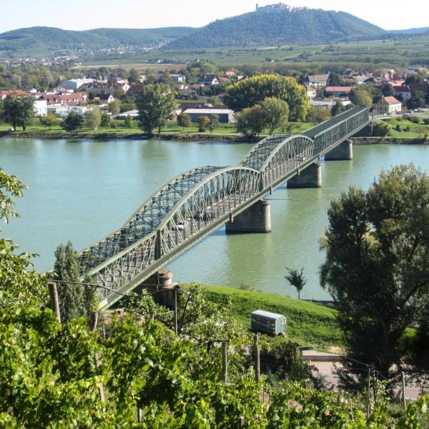 Eine grüne Stahlbrücke führt über einen breiten Fluss, im Hintergrund liegen Häuser und bewaldete Hügel.