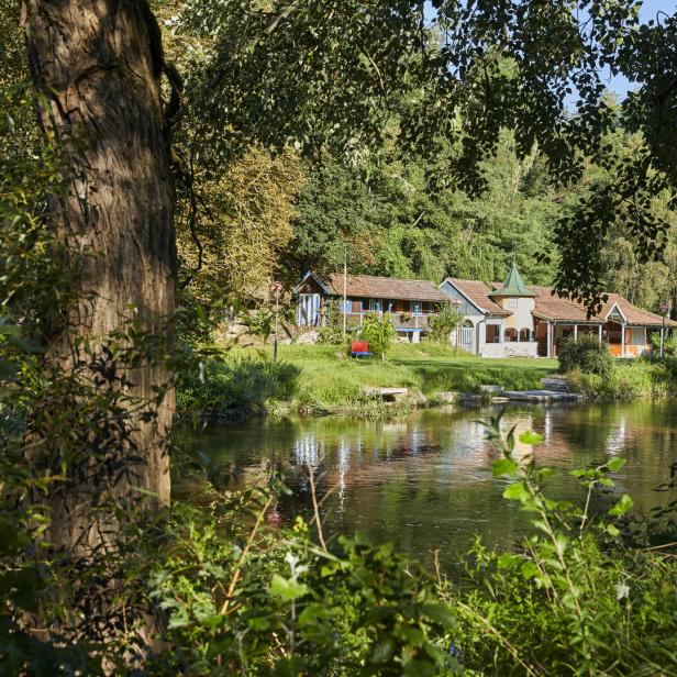 Grün-braunes Wasser, Bäume, eine Liegewiese und im Hintergrund ein Holzgebäude, das einst das Flussbad beherbergte.
