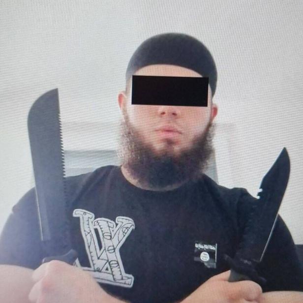 Ein Mann mit Bart und Mütze hält zwei große Messer vor der Brust.