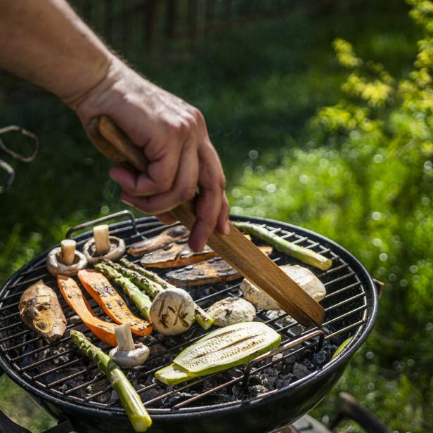 Kleiner Griller, auf dem Rost liegen Zucchini, Champignons, Spargel und Melanzani, ein Mann wendet das Gemüse mit einer Grillzange.
