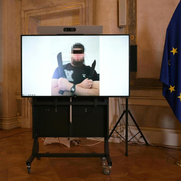 Auf einem Bildschirm ist ein Mann mit zwei Messern zu sehen; im Hintergrund die Europa- und Österreich-Flagge.