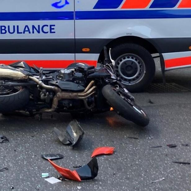 Umgekippte Motorrad-Unfallstelle vor einem Krankenwagen mit Trümmern auf der Straße.