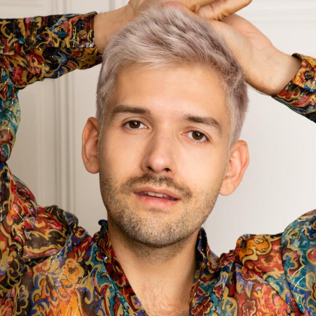 Ein Mann mit blond gefärbten Haaren und einem bunten Hemd posiert vor einer weißen Wand.