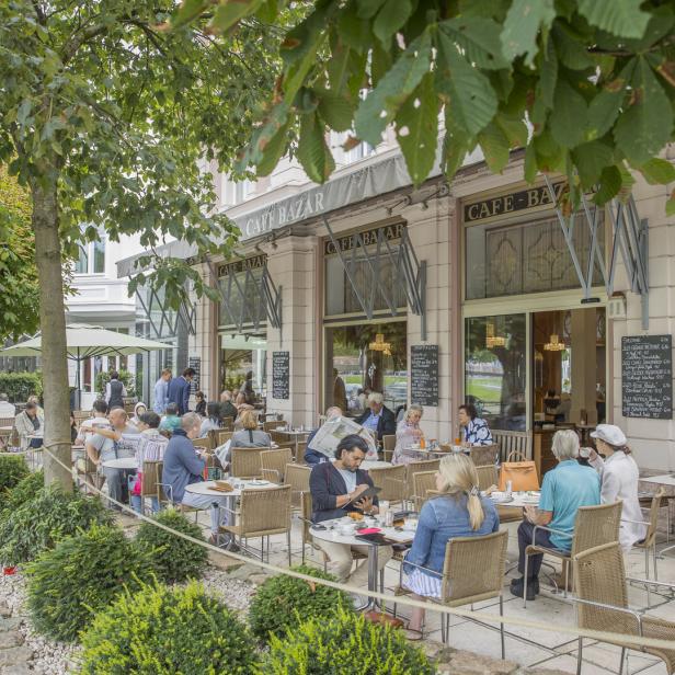 Gäste sitzen im Außenbereich des „Cafe Bazar“ unter schattigen Bäumen.
