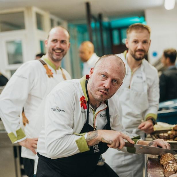 Ein Koch schneidet Fleisch in einer Restaurantküche, während andere Köche im Hintergrund stehen.