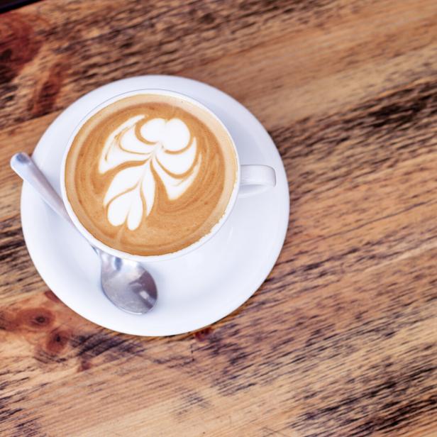 Ein Cappuccino mit Latte Art und ein pinkfarbenes Notizbuch auf einem Holztisch.