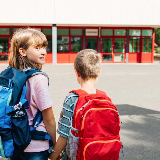 Zwei Kinder mit Rucksäcken halten Händchen vor einem Schulgebäude.