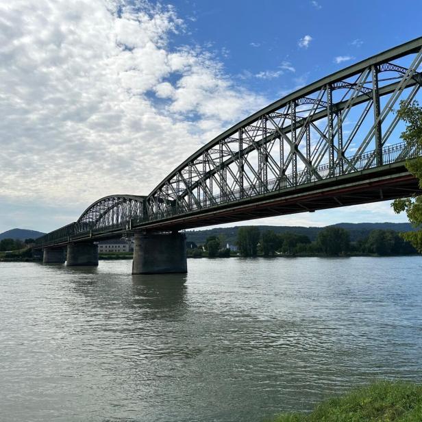 Eine grüne Stahlbrücke überspannt einen Fluss unter einem bewölkten Himmel.