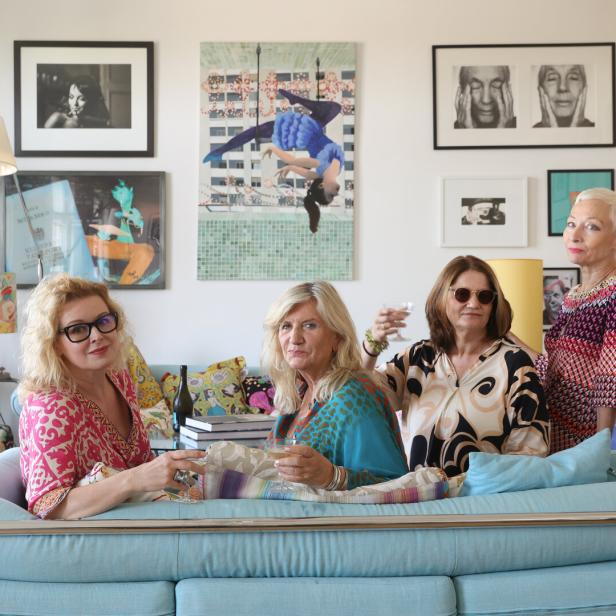 Vier Frauen sitzen auf einem Sofa und halten Gläser in einem Wohnzimmer mit Kunst an den Wänden.