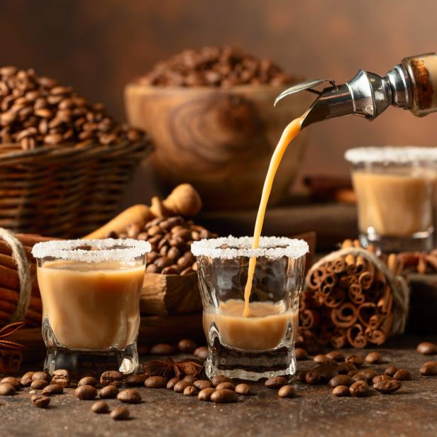 Ein Getränk wird in ein Glas mit Zuckerrand gegossen, umgeben von Kaffeebohnen und Zimtstangen.