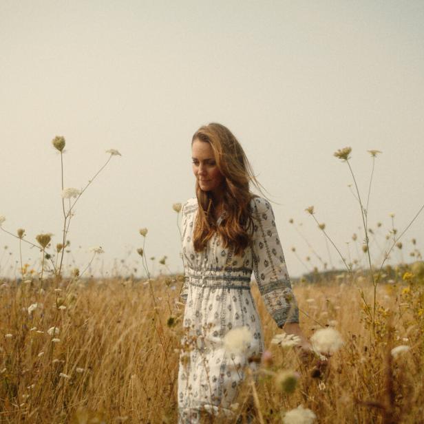 Eine Frau mit langem Haar geht durch ein Feld mit hohem Gras und Wildblumen.