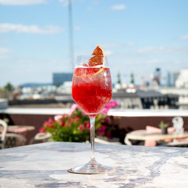 Ein roter Cocktail mit einer Orangenscheibe auf einem Tisch im Freien mit Blick auf die Stadt.
