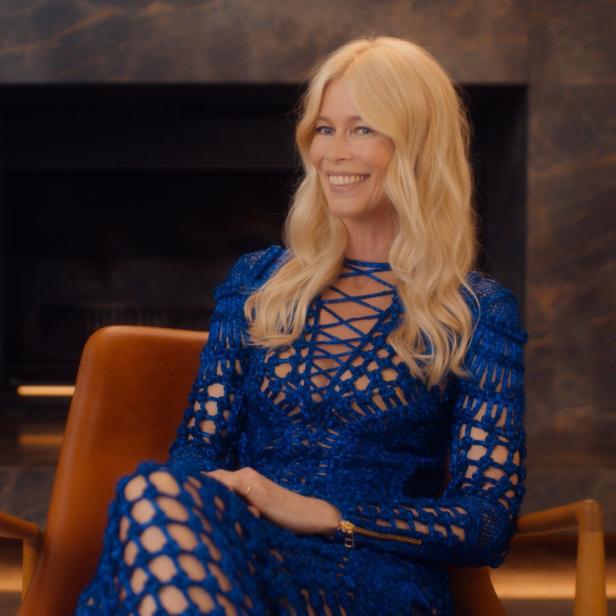 Claudia Schiffer sitzt in einem blauen Kleid in einem Sessel und lächelt.