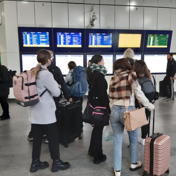 Reisende warten mit Gepäck auf einem Bahnhof vor Anzeigetafeln.
