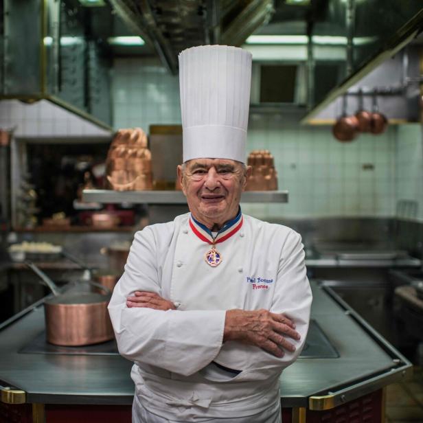 Paul Bocuse, ein Koch mit Kochmütze und Orden, steht in einer Restaurantküche.