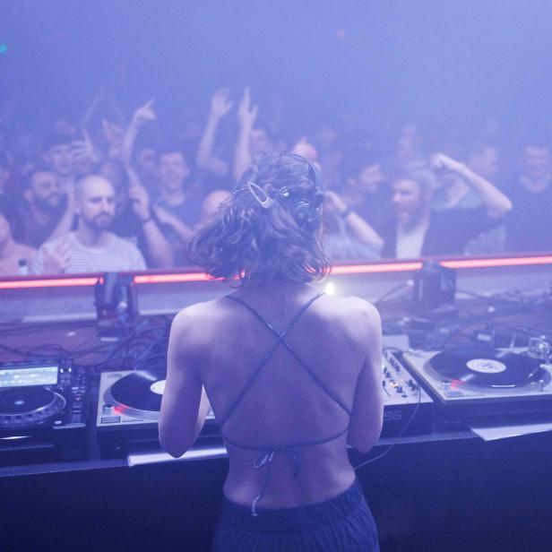 Eine junge Frau legt als DJ Platten in einem Techno-Club auf – von hinten fotografiert. Vor ihr jubeln Menschen.