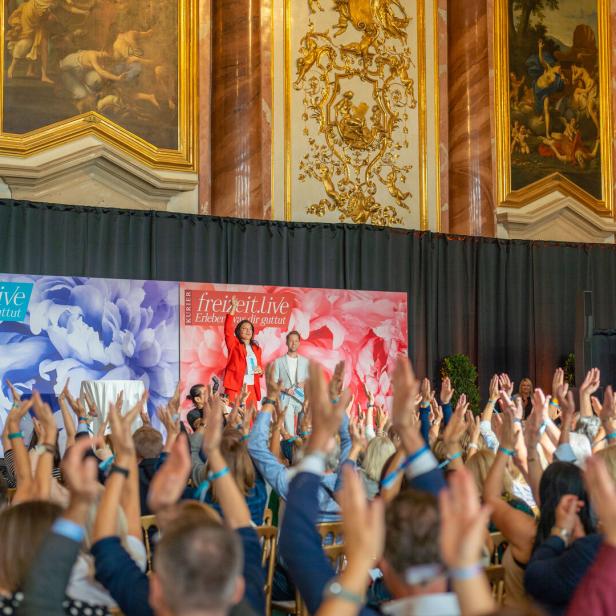 Ein barocker Festsaal mit vielen Besuchern, die die Hände in die Höhe reißen.