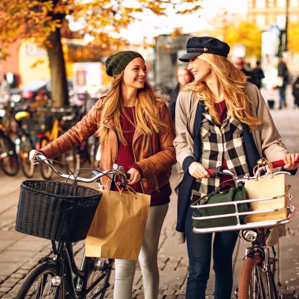 Zwei junge Frauen schieben ihre Fahrräder auf einem Bürgersteig im Herbst.