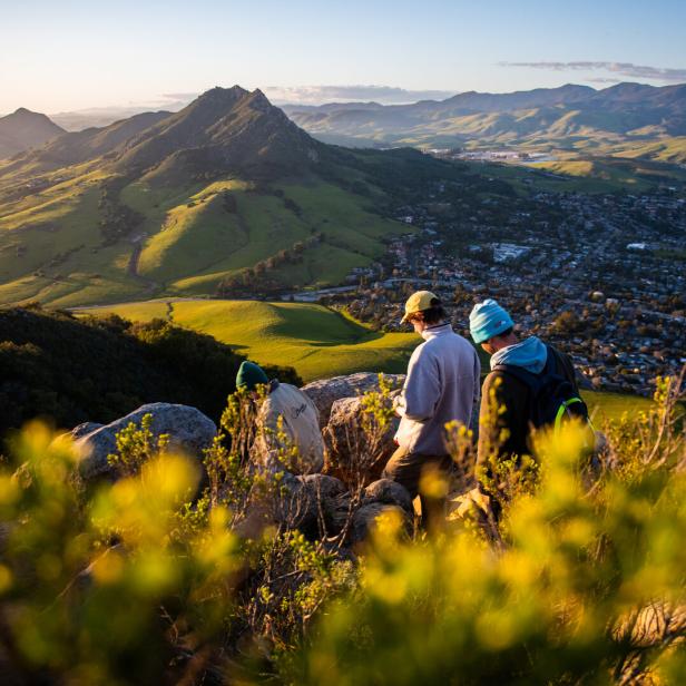 Eine Gruppe von Wanderern genießt die Aussicht auf eine Stadt und grüne Hügel im goldenen Abendlicht.