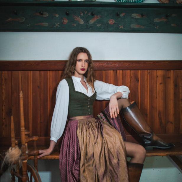 Eine Frau in einem Lena Hoschek Dirndl sitzt auf einer Holzbank neben einem Spinnrad.