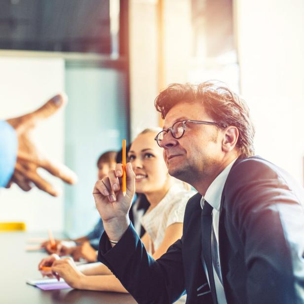 Ein Mann im Anzug hält einen Bleistift in die Höhe, während er in einer Besprechung sitzt.