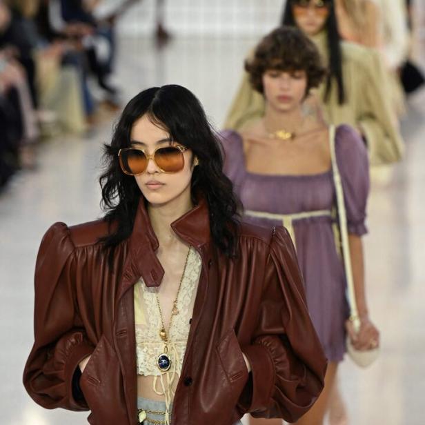 Eine Frau mit Sonnenbrille und brauner Lederjacke läuft auf einer Modenschau.