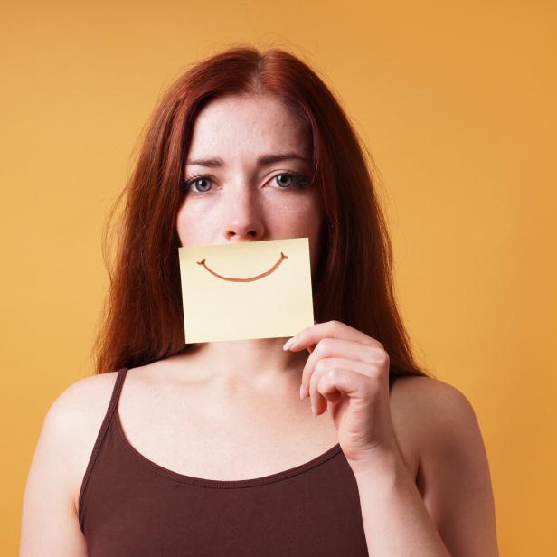 Eine Frau hält sich ein Post-It mit einem aufgemalten Lächeln vor ihren Mund..