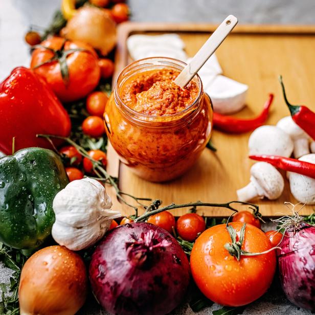 Ein Glas Ajvar umgeben von frischem Gemüse wie Paprika, Tomaten und Zwiebeln.