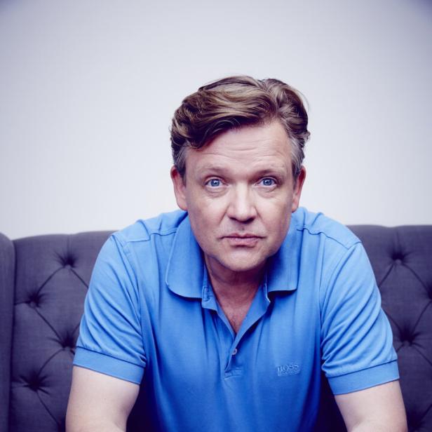 Ein Mann mit blauem Poloshirt sitzt auf einem grauen Sofa.
