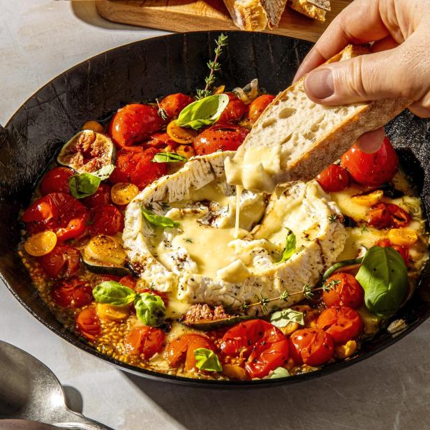 Ein Stück Brot wird in einen gebackenen Camembert mit Tomaten getunkt.