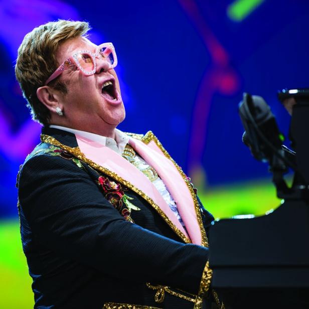 Elton John spielt Klavier und singt mit offenem Mund auf der Bühne.