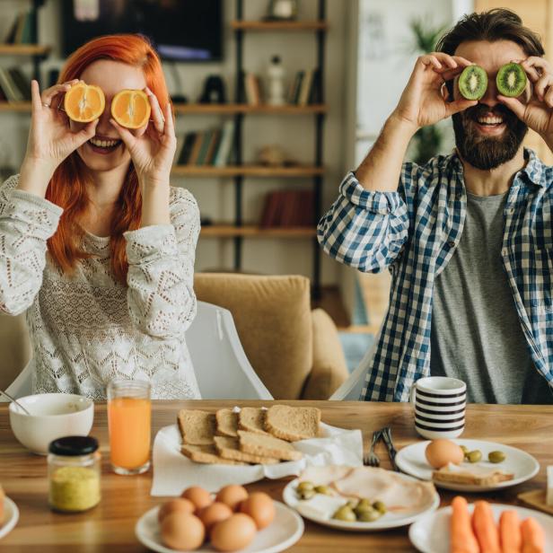 Ein Mann und eine Frau sitzen beim Frühstück und halten sich Orangen- und Kiwihälften vor die Augen.