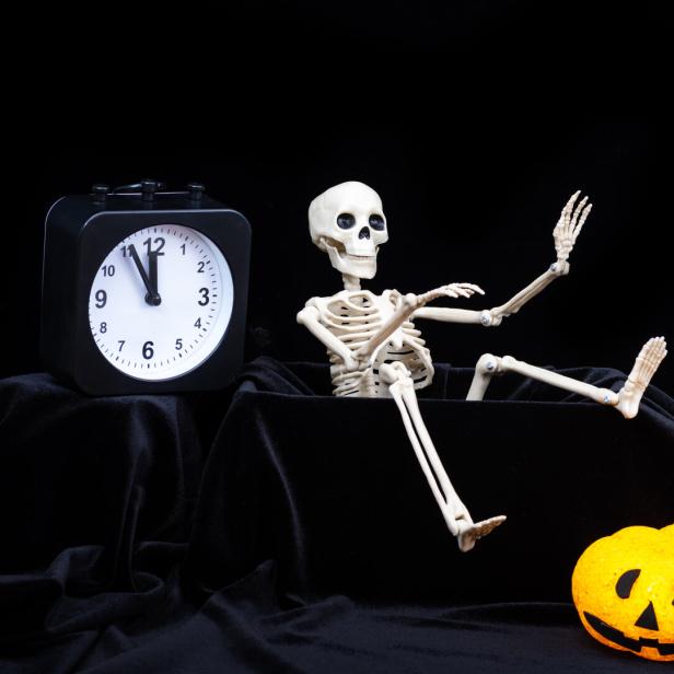 Ein Skelett sitzt neben einer leuchtenden Kürbis-Dekoration und einer Uhr, die fast Mitternacht zeigt.