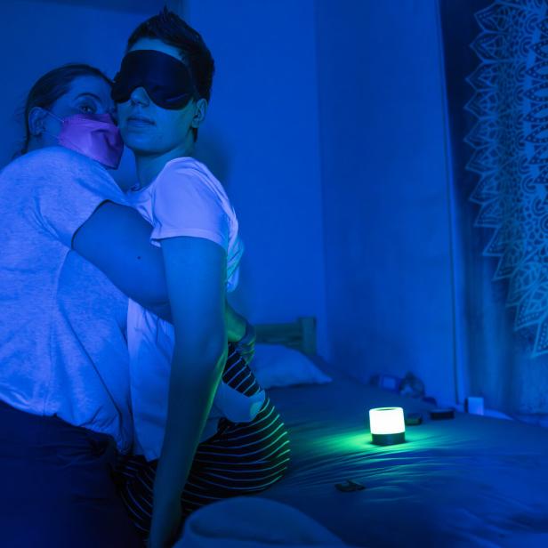 Eine Frau mit Maske umarmt eine Frau mit Schlafmaske in einem blau beleuchteten Raum.