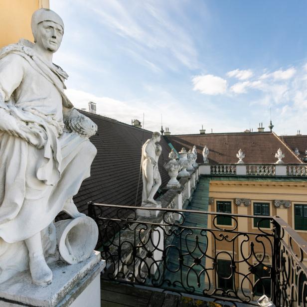 Steinfiguren schauen beim Balkon unterhalb der großen Uhr von oben auf den Ehrenhof des Schlosses Schönbrunn herab. Vor dem Balkon befindet sich ein Schmiedeisengitter, vor dem Dach eine steinerne Brüstung.