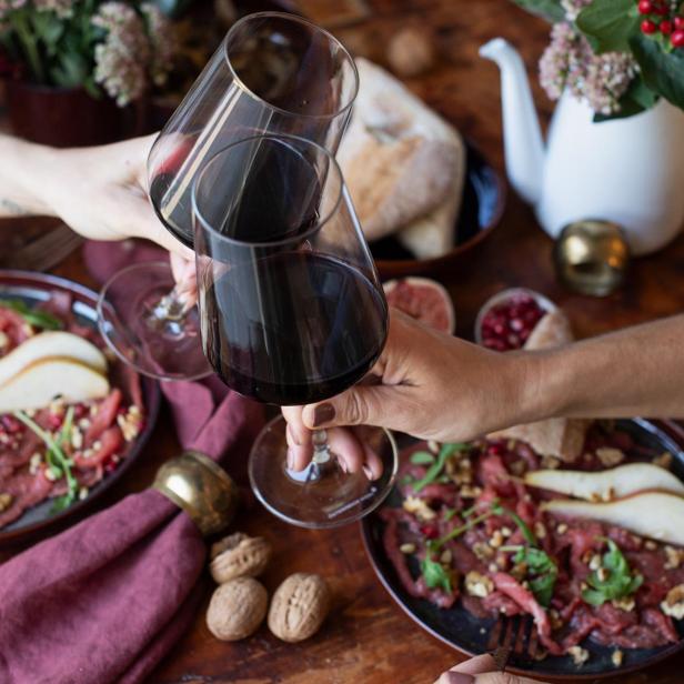 Zwei Personen stoßen mit Rotwein an. Auf dem Tisch befinden sich zwei Teller mit Essen.