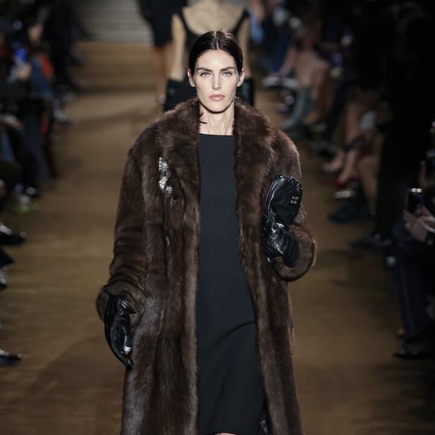 Eine Frau in einem braunen Pelzmantel und schwarzem Kleid läuft über einen Laufsteg.