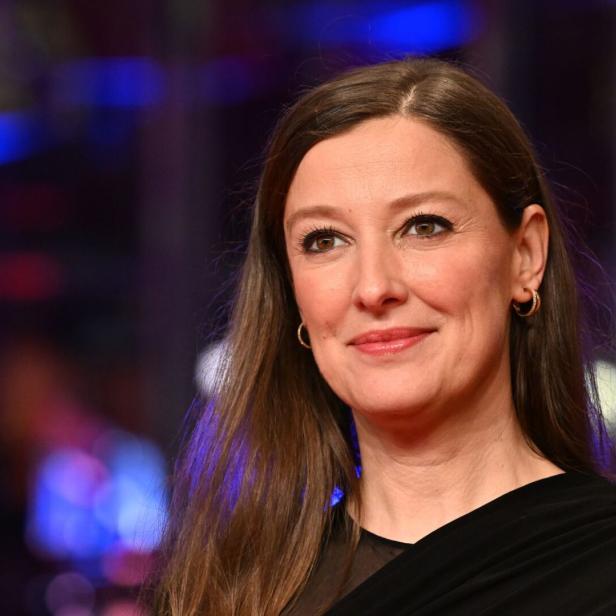 Alexandra Maria Lara mit braunen Haaren und Ohrringen lächelt vor einem verschwommenen Hintergrund.