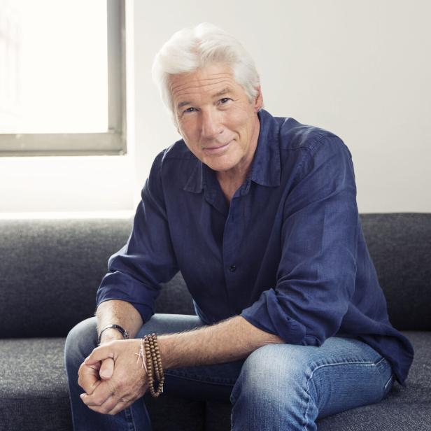 Richard Gere sitzt auf einer grauen Couch vor einem Fenster.