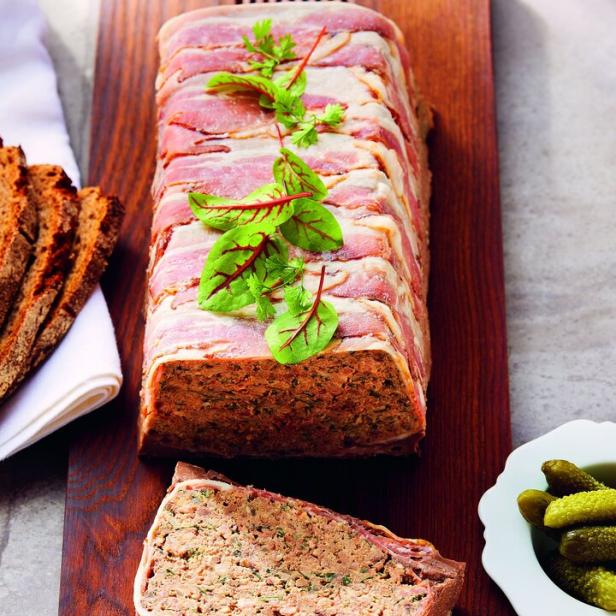Eine mit Speck umwickelte Hühnerleberterrine auf einem Holzbrett mit Brot und Gurken.