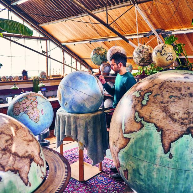 Ein Mann bemalt in einem Atelier einen Globus, umgeben von weiteren Globen unterschiedlicher Größe.