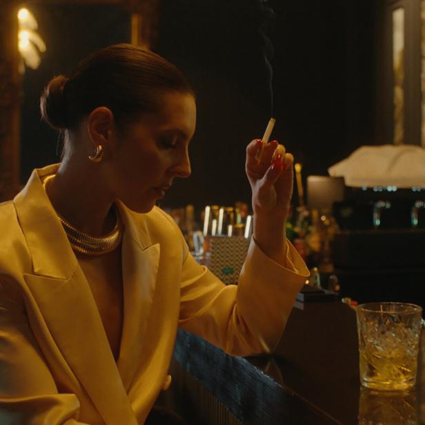 Eine Frau im gelben Blazer raucht an einer Bar.